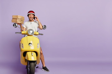 Friendly polite delivery man brings best restaurant food, checks order via smart phone, communicates with customer, tries to project positive image, carries pizza boxes, poses on bike over purple wall