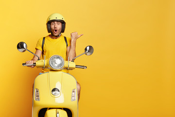 Impressed driver covers distance on yellow motorbike, wears helmet, indicates with great wonder aside, stops on road, demonstrates blank space for your advertising content, tries new vehicle. - obrazy, fototapety, plakaty
