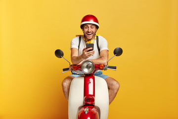 Joyful unshaven man sits on moped, wears red headgear, uses smartphone for communication on navigation, types message in motorbike service, happy to solve problem. Driving, technology concept