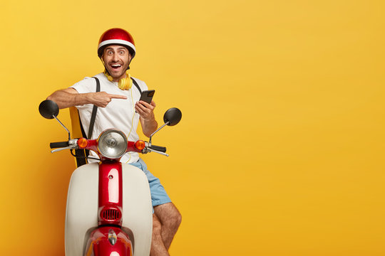 Isolated shot of happy delivery man points at smartphone used for finding route or right address, contacts with customer, poses with rucksack on motorbike, blank copy space on yellow background