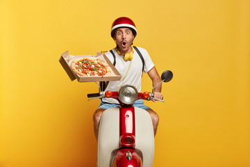 Stupefied male expert driver holds pizza box, delivers order to public, being on way to customer, shocked be late, poses on motorcycle in headgear, isolated on yellow background. Good service