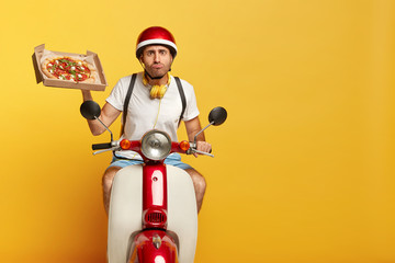 Purposeful delivery man focused into distance, rides on neccessary address to customer home, holds fresh baked pizza in box, wears casual apparel, has serious look, presses lips together. Shipping