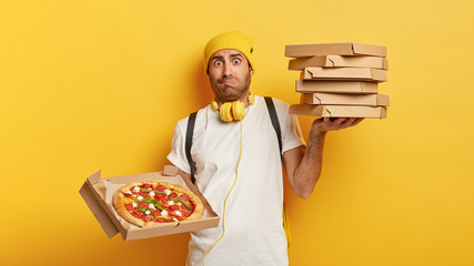Photo of busy delivery man carries stack of heavy boxes filled with pizza, wears yellow hat and white t shirt, busy delivering fast food, has dissatisfied facial expression. Online order and service