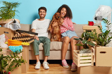 Relocation, renovation and removal concept. Happy diverse couple sit on sofa with favourite dog, have break on moving day, busy unpacking, have mess in new flat, many packages, remove to own house