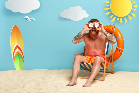 Handsome redhead man rest at sea resort, keeps coconuts near eyes as binoculars, gazes into distance, wears shorts, sits in comfortable beach chair near lifering, surfboard. Good summer weather