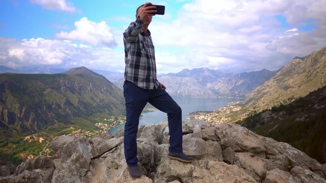 Middle-Aged Man Taking Selfies on Smartphone on Top of Mountain. Beautiful Landscape with Lake and Mountains on Background.