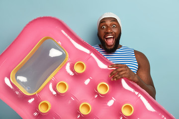 Happy overjoyed surprised man with dark skin gazes at camera with great interest and excitement, wears rubber swimhat, carries pink mattress on beach, has fun during recreation time. Summer concept