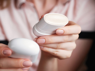 Woman's beauty blogger hands holding opened professional white glossy cosmetics sick with sample trendy tone for shiny skin with blurred body background warm cozy tones and copyspace