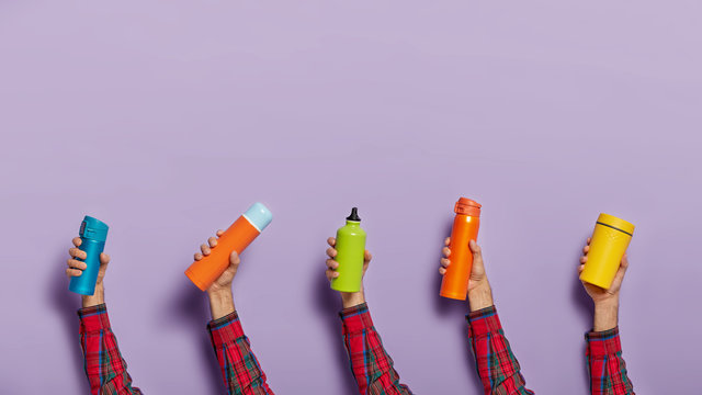 Horizontal shot of unknown person holds various flasks or termo mugs of different colors, filled with hot beverage, isolated on purple background. Close up. Beverage in man hands. Containers for drink