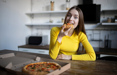 Funny brunette girl in yellow sweater eating pizza at kitchen