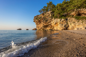 Fototapeta na wymiar Sea bay with rocky cliff covered with growing green pine trees. Charming splashing water at seashore of empty beach in Turkey. Horizontal color photography.