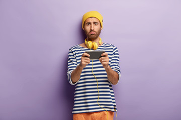 Unhappy teenager holds modern cell phone device, looks sadly at camera, lost online game, messages via application, listens radio music in headphones, cannot install new app. Sad blogger indoor