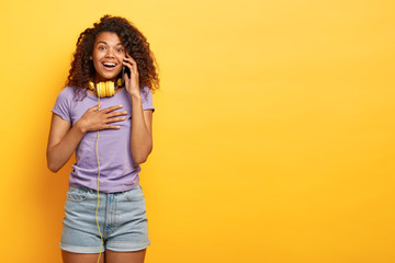 Positive young female with Afro hairstyle, phones to best friend, uses free internet connection, gazes happily at camera, wears casual violet t shirt and denim shorts, has pleasant conversation