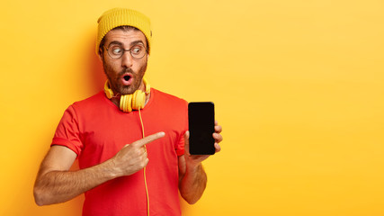 Stupefied sales manager demonstrates new smartphone, surprised with new functions, points at mock up screen for your promotional content, wears bright yellow hat and red t shirt. Technology retail