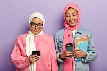 Two happy Muslim students have coffee break, chat online on smart phone, wear veils, stand closely against purple background, hold spiral notebook, diary and textbook, study together at college