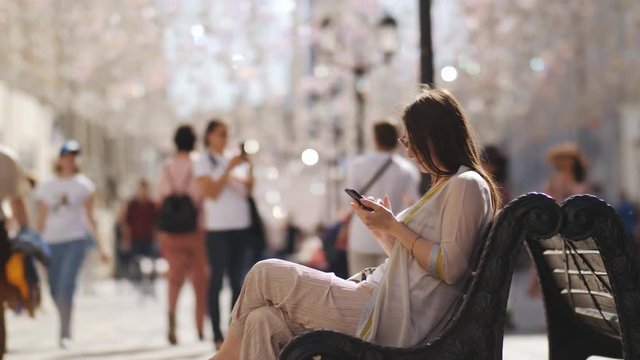Young beautiful woman using a smartphone sitting on bench on the street in the city centre, in the background, and the foreground people walking and going about their business.