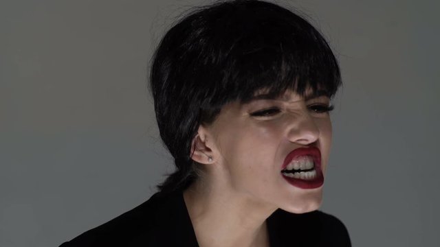 Young woman with red lips desperate screaming. Evil woman with black hair and red lips. Crazy woman are scary and insane. Red lipstick and white teeth. Screaming woman over gray background.