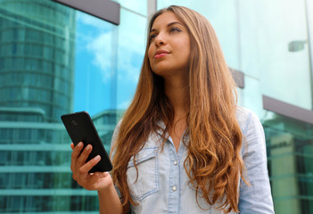 Young serious and determined business woman standing in front of modern building with phone in her...