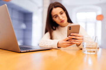 Beautiful young modern casual woman is using laptop and phone on kitchen at home. Working online.