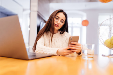 Beautiful young modern casual woman is using laptop and phone on kitchen at home. Working online.