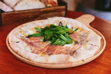 Parma Ham Pizza topping with rocket on rounded wooden plate with sliced bread in wooden box in the background.