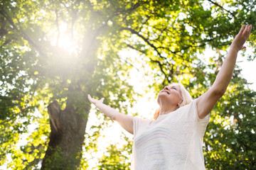 Senior scandinavian woman by the tree holding hands up. Back view