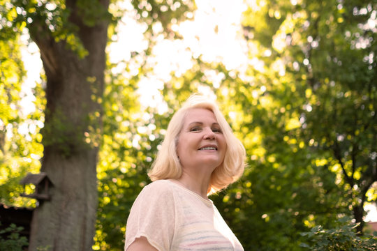 Senior scandinavian woman in summer garden between trees smiling looking at camera.