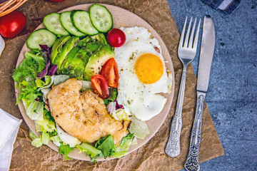 Served healthy breakfast with fried eggs and vegetable Top view of plate of tasty fried eggs with chicken and avocado with colorful vegetable salad placed next to knife and fork on kitchen table