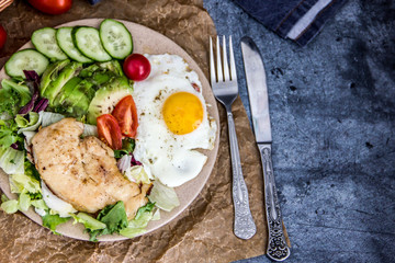 Served healthy breakfast with fried eggs and vegetable Top view of plate of tasty fried eggs with chicken and avocado with colorful vegetable salad placed next to knife and fork on kitchen table