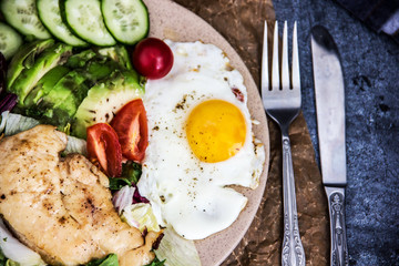 Served healthy breakfast with fried eggs and vegetable Top view of plate of tasty fried eggs with chicken and avocado with colorful vegetable salad placed next to knife and fork on kitchen table
