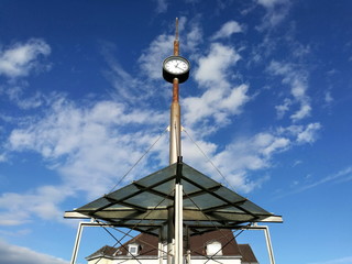 Überdachter Wartebereich mit alles überragender Bahnhofsuhr vor blauem Himmel mit weißen Wolken...