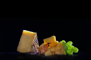 Pieces of cheese on a black background with grapes. Copy space. Still life on a black background. Homemade cheese concept.