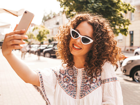 Beautiful smiling model with afro curls hairstyle dressed in summer hipster white dress.Sexy carefree girl posing in the street in sunglasses.Taking selfie self portrait photos on smartphone