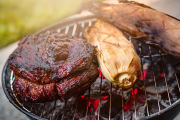 A large beef brisket and corn in the husk being barbequed on a small charcoal hibachi grill with glowing coals underneath with a sunburst.
