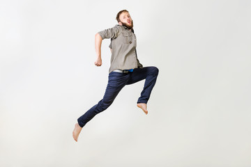 Full-length photo of beard man in casual t-shirt and jeans running or jumping in air makes funny faces over gray background