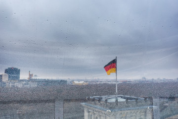 Blurred German flag and urban skyline viewed through a window full of raindrops on a rainy and cloudy day. Focused on the front, the window. Copy space.