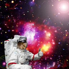 Astronaut gives thumbs-up against outer space, galaxies and stars. The elements of this image...