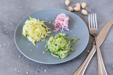 crushed cucumbers, zucchini, radish on a gray plate on a gray concrete background, fork, knife,