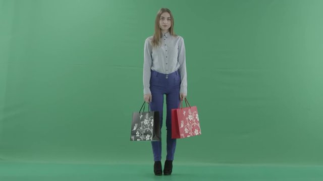 Woman with her purchases is posing for camera. Lady with straight brunette hair is wearing casual shirt and jeans. Female shopper holding shopping bags is standing on green background in a studio.