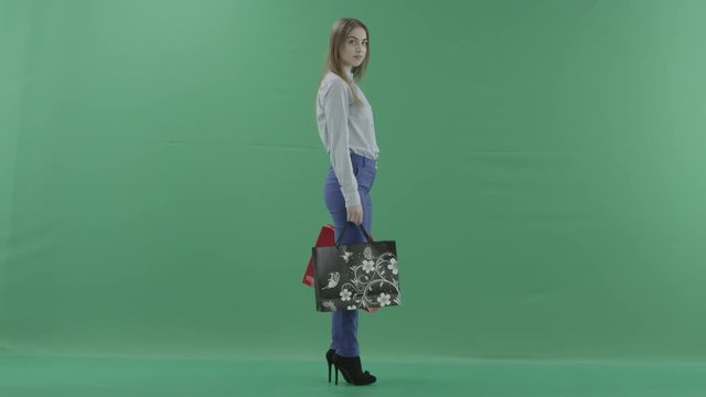 Woman with her purchases is posing for camera. Lady with straight brunette hair is wearing casual shirt and jeans. Female shopper holding shopping bags is standing on green background in a studio.