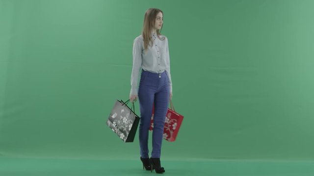 Woman with her purchases is posing for camera. Lady with straight brunette hair is wearing casual shirt and jeans. Female shopper holding shopping bags is standing on green background in a studio.