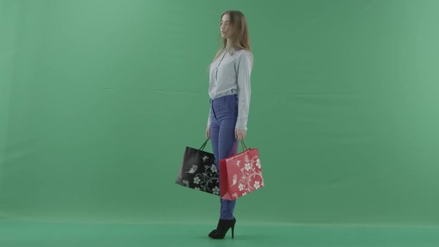 Woman with her purchases is posing for camera. Lady with straight brunette hair is wearing casual shirt and jeans. Female shopper holding shopping bags is standing on green background in a studio.