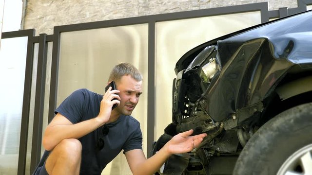 car accident concept. man in a state of shock talking on the phone after a car accident, standing by a car with a broken bumper