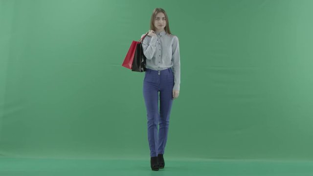 Woman with her purchases is posing for camera. Lady with straight brunette hair is wearing casual shirt and jeans. Female shopper holding shopping bags is standing on green background in a studio.