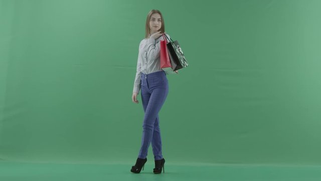 Woman with her purchases is posing for camera. Lady with straight brunette hair is wearing casual shirt and jeans. Female shopper holding shopping bags is standing on green background in a studio.
