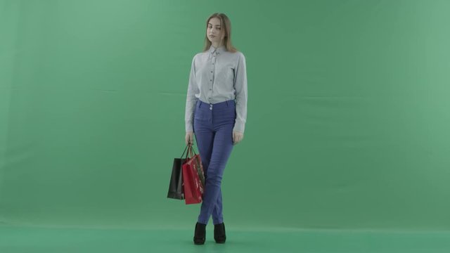 Woman with her purchases is posing for camera. Lady with straight brunette hair is wearing casual shirt and jeans. Female shopper holding shopping bags is standing on green background in a studio.