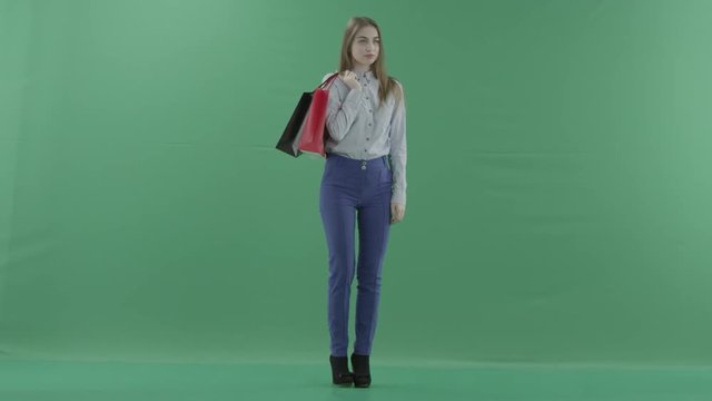 Woman with her purchases is posing for camera. Lady with straight brunette hair is wearing casual shirt and jeans. Female shopper holding shopping bags is standing on green background in a studio.
