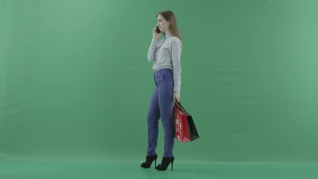 Woman with her purchases is talking on her mobile phone. Lady with straight brunette hair is wearing casual shirt and jeans. Female shopper holding shopping bags is standing on green background in a