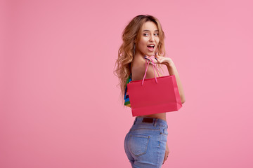 Portrait of an excited beautiful smiling girl wearing colorful clothes holding shopping bag isolated over pink background. Place to sign or text on the package