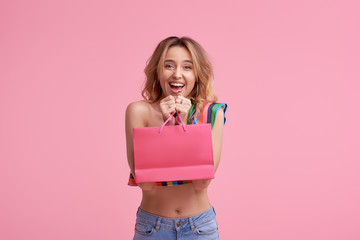 Portrait of an excited beautiful smiling girl wearing colorful clothes holding shopping bag isolated over pink background. Place to sign or text on the package